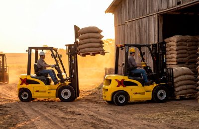 Duas empilhadeiras Techlift trabalhando em um campo carregando sacos de safra para o galpão.
